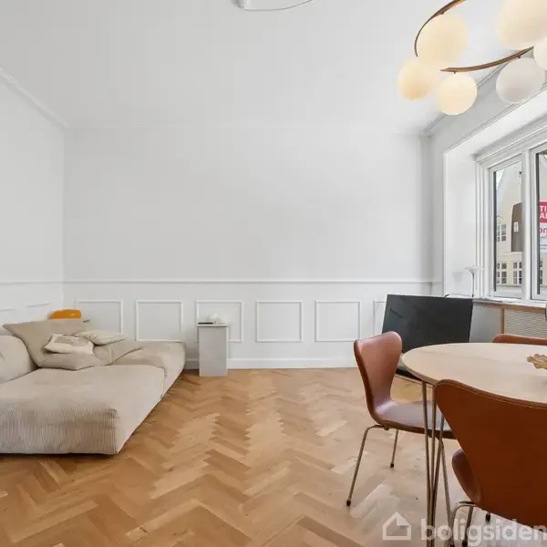 En beige sofa står langs en hvid væg med paneler. Et rundt bord med brunlige stole er foran et stort vindue. Udenfor ses bygninger og et skilt med teksten "Til Salg".