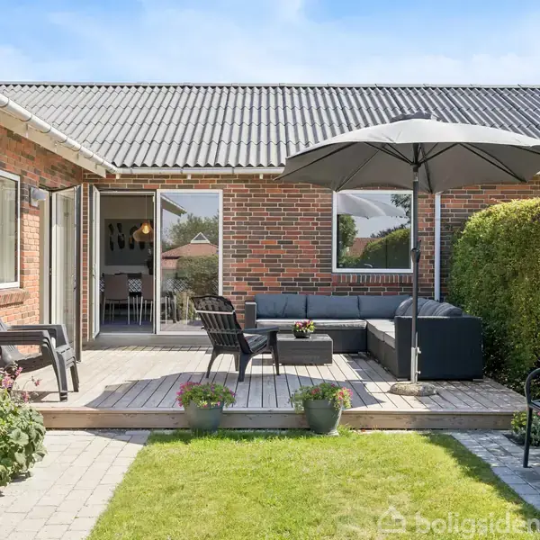 Terrasse med sort sofa, bord og parasol. Fliser og græs omgiver området. Murstenshus i baggrunden med åben dør til indendørs spisestue. Hæk til højre, blomsterkrukker på terrassen.