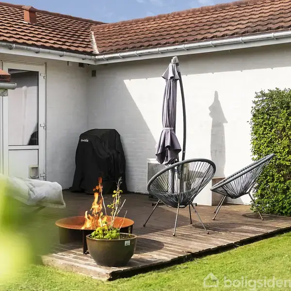 Terrasse med to stole står på et trædæk. En grill er dækket af et overtræk ved siden af. En parasol er klappet sammen. En ild brænder i en ildskål på det grønne græs.