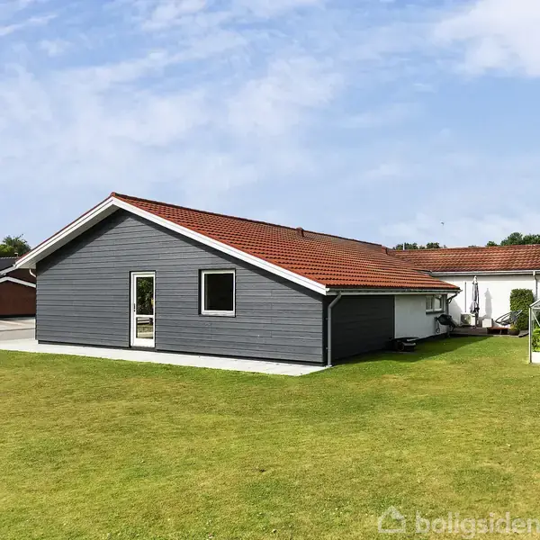 Grå hus med rødt tegltag står i en velplejet have med en græsplæne. Ved siden af huset er et drivhus. Skyfri blå himmel i baggrunden.