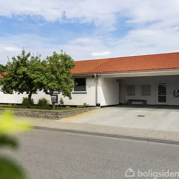 Huset har et hvidt ydre og rødt tegltag, carport med en bænk, omgivet af en have med et træ, beliggende ved en stille gade.