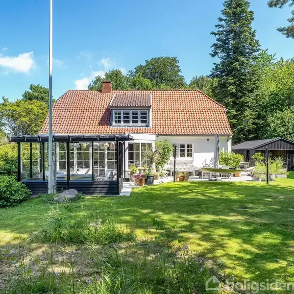 Huset står i en grøn have med træer, udstyret med en terrasse og flagstang. Det er en villa med rødt tegltag og har store vinduer og et udestueområde.