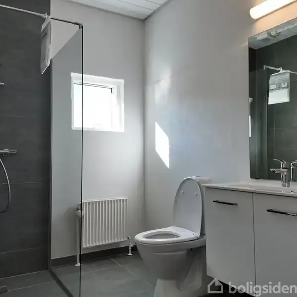 Badeværelse med bruser bag glasvæg, toilet og vask med spejl i moderne stil. Lysegrå fliser og hvidmalede vægge skaber et lyst, minimalistisk rum. Lille vindue kaster naturligt lys ind.
