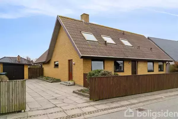 Et gult murstenshus med skråt tag og tre ovenlysvinduer står ved en flisebelagt indkørsel. Huset er omgivet af et brunt plankeværk i et roligt boligområde.