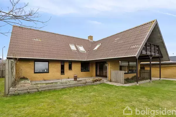 Et gult murstenshus med store skråtag og flere vinduer. Huset står på en grøn græsplæne. Der er terrasse på siden under et overdækket område.