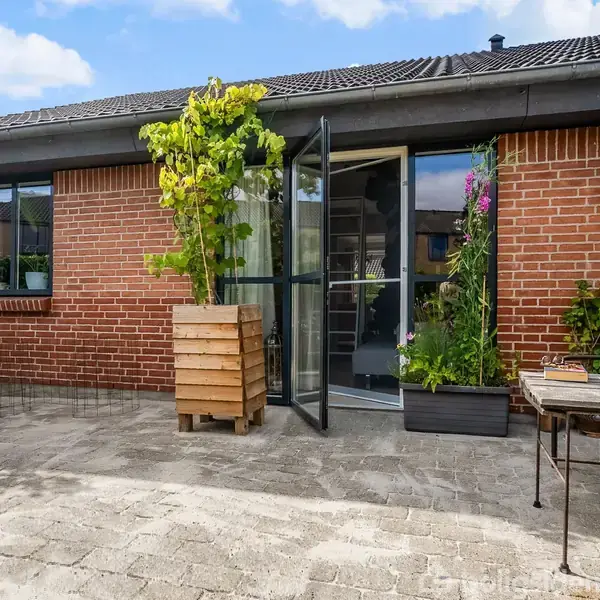 En terrasse i mursten med grønne planter i krukker foran et hus. En glasdør står åben, og der er møbler ved siden af, mens himlen er klar og blå.
