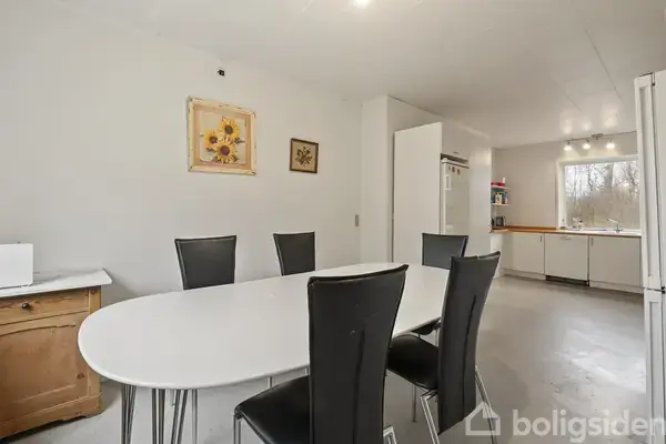 Ovalt spisebord med seks sorte stole står i et lyst køkken med hvide skabe. Et lille skab står til venstre, og to malerier hænger på væggen.