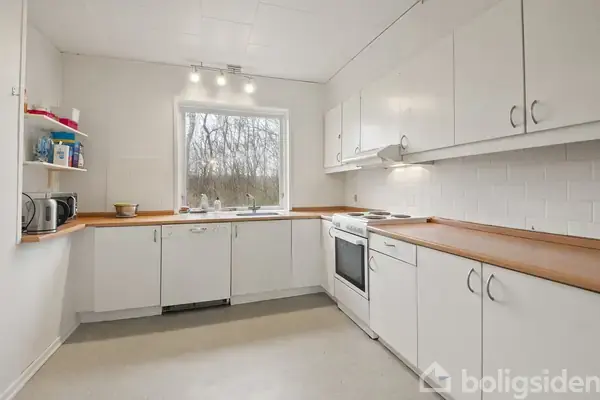 Et hvidt køkken med træbordplader og flere skabe uden greb. Et vindue giver udsigt til træer udenfor. En komfur og vask er placeret langs væggen.