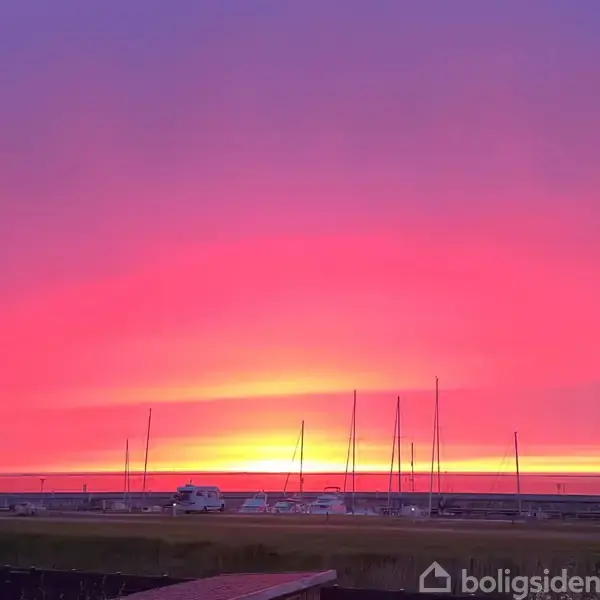 Solnedgang med intense lyserøde og gule nuancer. Enkelte både og master står stille ved kysten. I forgrunden ses biler og en bygning med en klar himmel ovenfor.