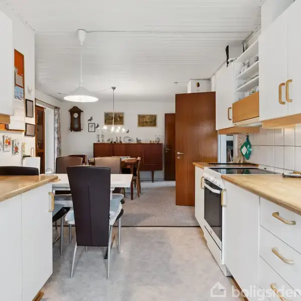 Et køkken med hvide skabe og træbordplader. En mikroovn står på bordet. I baggrunden ses et spiseområde med en rektangulær bord og stole samt vægkunst og en dør.