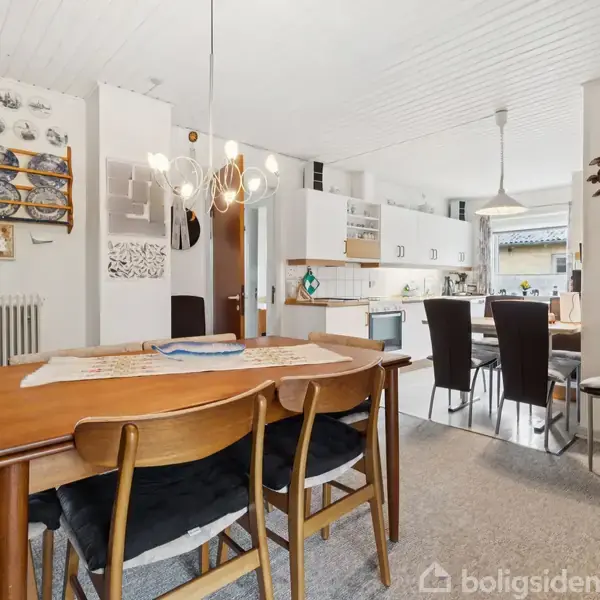 Et spisebord med træstole står i en spisestue. Væggene har tallerkendekorationer og hylder. Baggrunden viser et køkken med lyselementer og spisebord med mørke stole.