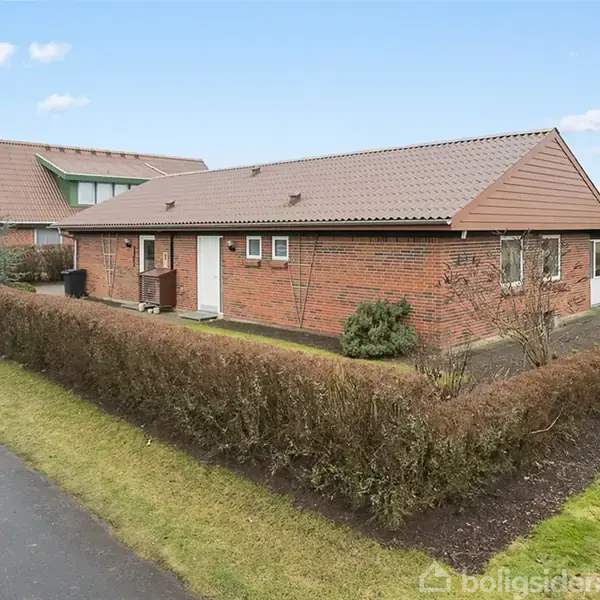 En murstensvilla med rødt tag står på en hjørnegrund omgivet af en lav hæk. Villaen har hvide døre og vinduer. Brolagt indkørsel og græsplæne ses omkring huset.