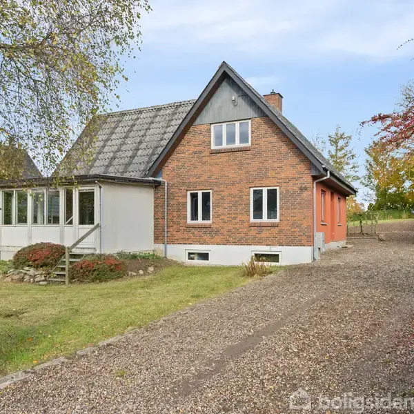 Et rødstenshus står stille omgivet af en grusvej og græsplæne. Ved siden af huset ses en indgang med vinduer, mens træer omgiver ejendommen i en rolig, landlig kontekst.