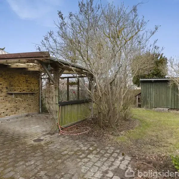 En gårdhave med en brolagt terrasse og en pergola dækket af klatreplanter. En brun murstensbygning med en grøn dør ses til venstre, omgivet af græs og træer.