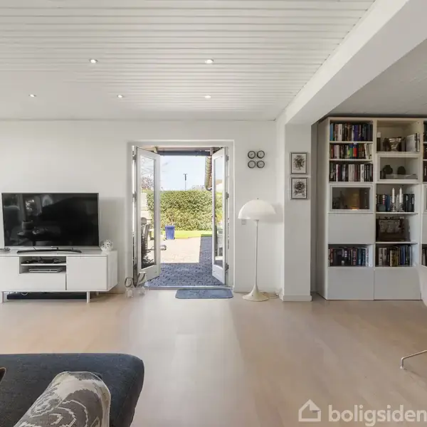 Stue med sofa, fjernsyn på tv-bord, og indbygget bogreol. Åben dør fører til have med grønne buske. Lampe og runde billeder på væggen. Lysindfald fra vindue.