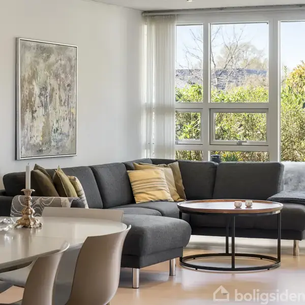 Sofa i hjørne fyldt med puder, står ved vinduer med udsigt til have. Rundt sofabord står foran. Elegant spisestue med blomster på bordet i forgrunden.