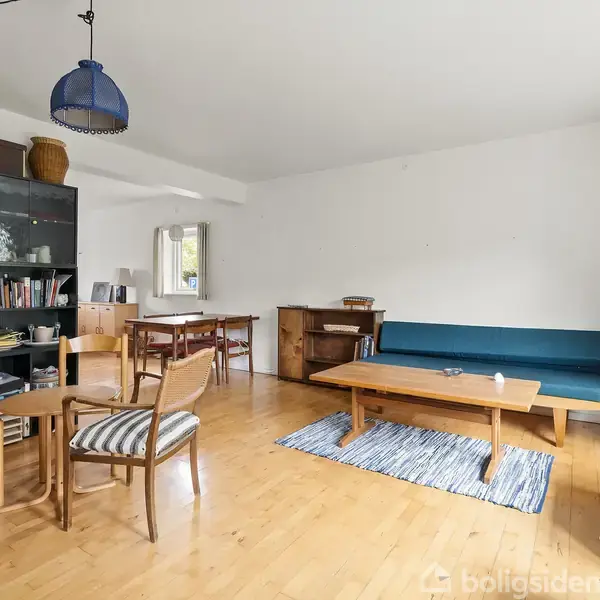 Stue med træmøbler, hvor en blå sofa står op ad en hvid væg. Et sofabord med en skål foran en fjernsyn. Reol med bøger og kurve i baggrunden. Ingen tekst.
