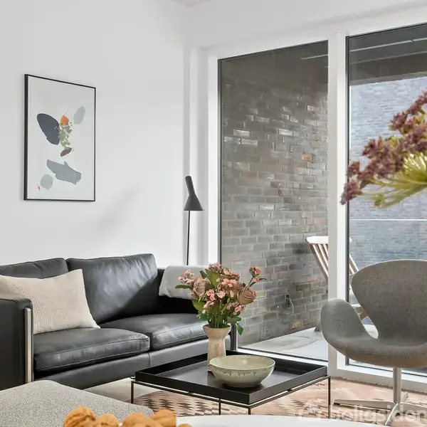 Sort sofa med puder står i moderne stue ved siden af et glasvindue. En plakat hænger på væggen, og en lampe står i hjørnet. Blomster er på sofabordet.