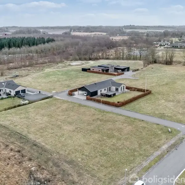 Et parcelhus ligger på en stor åben grund omkranset af en lav hæk. Det er omgivet af lignende huse og åbne grønne arealer i et landligt område.