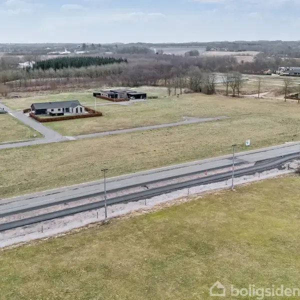 Åben landparcel med græs dominerer billedet, omkranset af asfaltvej og spredte moderne huse i baggrunden. Vejskilte og lysmaster til stede, med skov i horisonten.