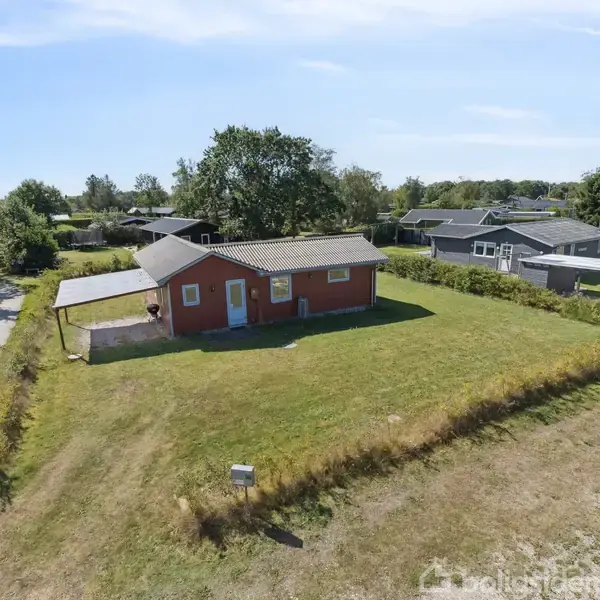 Et rødt hus med gråt tag står midt på en græsplæne. Ved siden af huset er en grusvej, og der er andre huse i baggrunden omgivet af træer og buske.