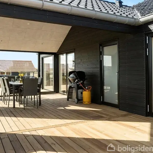 Bord og stole står på en træterrasse foran en sortmalet bygning. En grill med en gasflaske står ved siden af døren. Et tag skygger delvist området. Tekst: "Modelfoto".