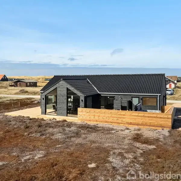 Et sortmalet sommerhus står ved en kystnær beliggenhed. Huset har en træterrasse foran og er omringet af åbne arealer og andre spredte huse.