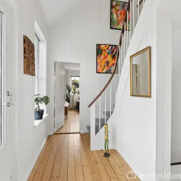 Trappeopgang med hvidmalet trævæg, et farverigt abstrakt maleri på væggen og en smal gang bagved. En træfigur står ved foden af trappen, og et stort vindue lader lys ind.