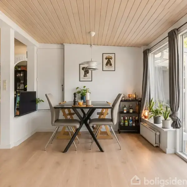 Et spisebord med fire stole står i en lys, moderne spisestue med trægulv og loft. Rummet indeholder planter, en reol og hængende billeder, med store vinduer mod en terrasse.