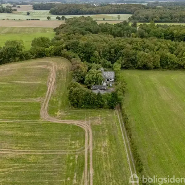 En bygning omgivet af træer står midt i åbne marker. En grusvej slanger sig gennem landskabet, som er domineret af grønne felter og en tæt skov horisonten.
