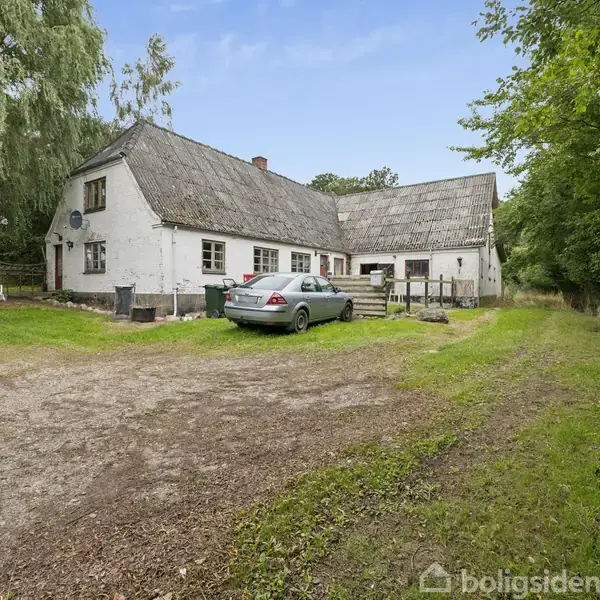 Huset, en traditionel bygning med stråtag og hvid facade, står i et grønt landskab. En bil holder parkeret foran på en jordvej omringet af træer og græs.