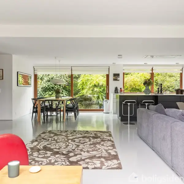 En moderne stue med grå sofa og rødt lænestol, beliggende foran en pejseindsats. Spisebord og stole opstillet nær store vinduer med udsigt til grøn have. Gulvtæppe med blomstermønster.
