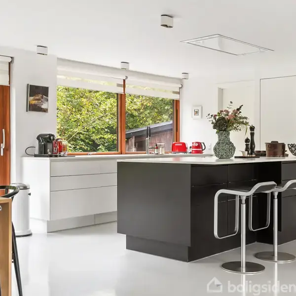 Et moderne køkken med en central sort ø, hvorpå der står en vase med blomster og andre køkkenartikler. Store vinduer omkranser rummet og giver udsigt til en grøn have.
