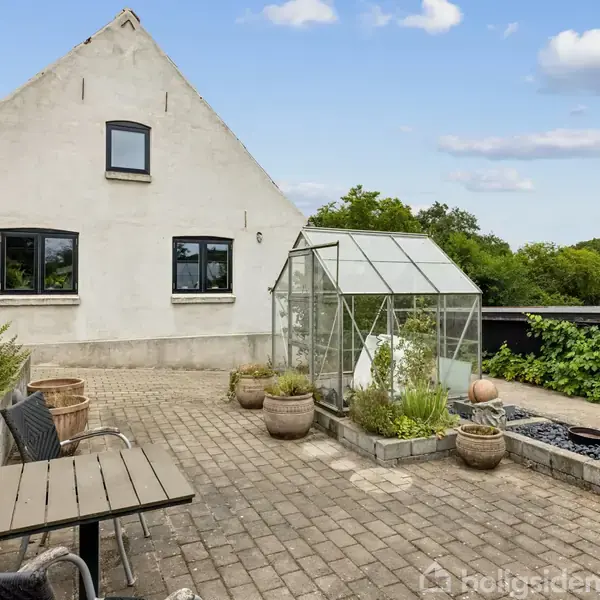 Et hvidt hus med tre vinduer står ved en flisebelagt terrasse. På terrassen er et lille drivhus omgivet af krukker. I baggrunden er frodigt grønt landskab.