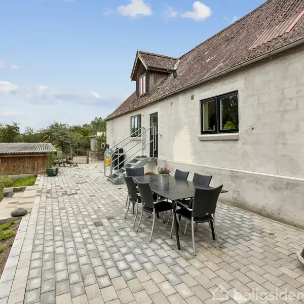 Et bord med stole står på en flisebelagt terrasse ved et hvidt hus med et skråtag. En parasol er ved siden af bordet. Græsplæne og træer omkranser området.