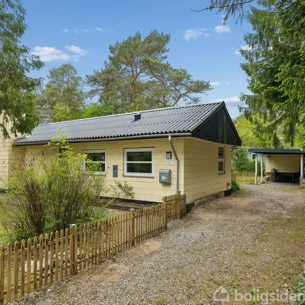 Et gult hus med sort tag står stille på en grusvej. Omgivet af høje træer og grønne buske i en skovklædt have. En carport ses i baggrunden.