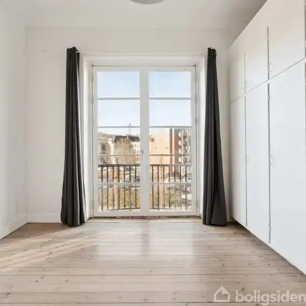 Et lyst værelse med trægulv og hvide vægge, handling: rummets store glasdøre åbner ud til en balkon med udsigt over bybebyggelse. Gardiner hænger ved døren, og indbyggede skabe ses til højre.