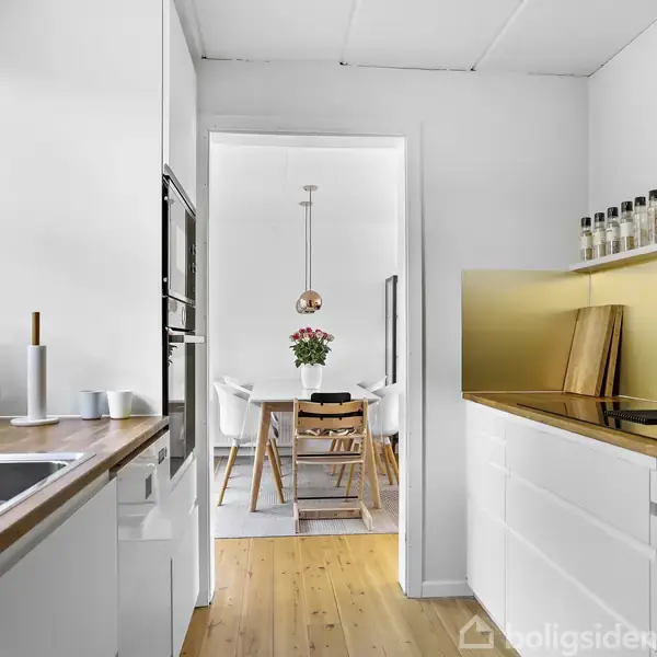 Køkken med hvide skabe og træbordplader, udstyret med vask og indbygget ovn. I baggrunden et spisested med bord, stole og blomster på bordet. Naturligt lys fra vinduet.