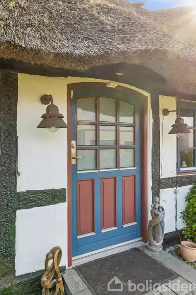 Dør med blå ramme og røde paneler står lukket; to lanterner hænger på væggen. Stråtag og traditionelle bindingsværk træpaneler danner en rustik indgang. Skulpturer flankerer trappen.