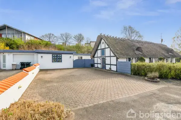 Bindingsværkshus med stråtag står stille på en brolagt gårdsplads omkranset af buske. I baggrunden ses en grøn bakke med enkelte træer under en klar blå himmel.