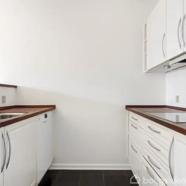 Hvidt køkken med træbordplader, står funktionelt opstillet langs væggene. Køkkenet har en vask, komfur og masser af skabe. Indretningen er minimalistisk med et lyst og rent udtryk.