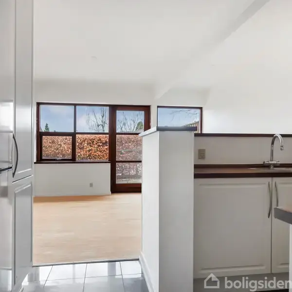 Køkken med hvidt interiør og stålgreb, indeholder en vask og bordplade. I baggrunden ses en stue med store vinduespartier og udsigt til en hæk.