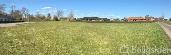 En stor grøn græsplæne strækker sig i forgrunden, omgivet af en vej og flere huse i baggrunden under en blå himmel med få skyer.