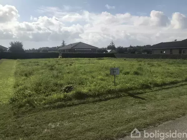 En græsmark med et skilt i forgrunden, hvor der står “11”. I baggrunden er der et hus omgivet af hække og andre bygninger under en delvist skyet himmel.
