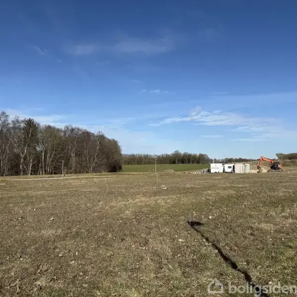 Gravearbejdsmaskine arbejder på felt i landligt område med åbne marker og træbevoksning i baggrunden under en blå himmel.