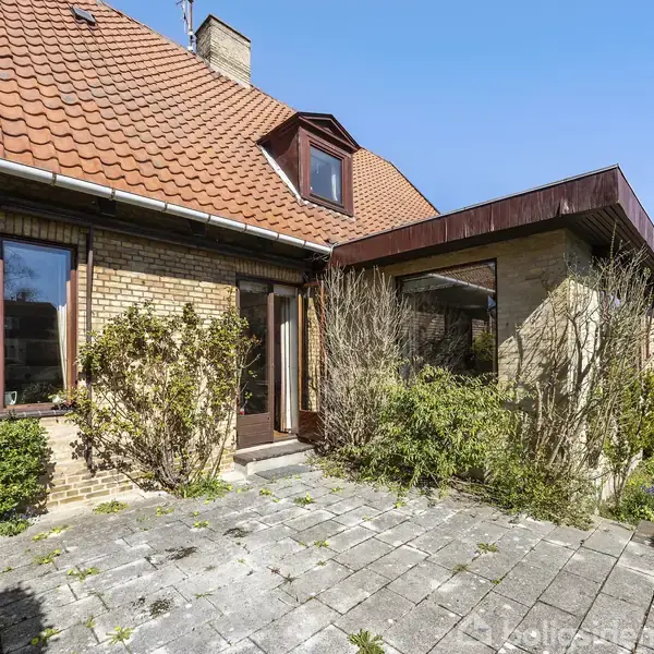 Rødstenshus med rødt tegltag, har en stor terrasse med flisebelægning. Omgivet af frodig have med buske og træer. Huset har store vinduer og en dør mod terrassen.