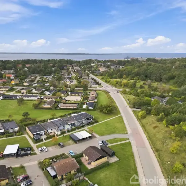 Luftfoto af et boligområde med rækkehuse omgivet af grønne marker og skov. En hovedvej løber gennem landskabet, og i baggrunden ses vand og horisonten.