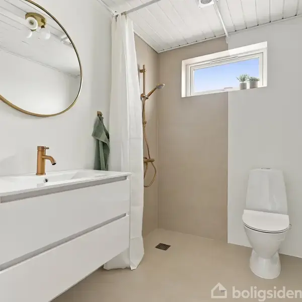 Badeværelse viser håndvask med rundt spejl og bruseområde med bruseforhæng. Et vindue over toilettet giver lys. Håndklæde hænger ved vasken, mens gulvet er lyst flisebelagt.