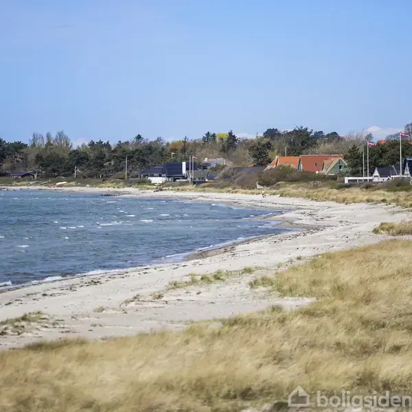 Strand med græs i forgrunden strækker sig langs kysten, mens bølgerne ruller ind. Huse og træer ses i baggrunden under en klar, blå himmel. En vindmølle står i horisonten.