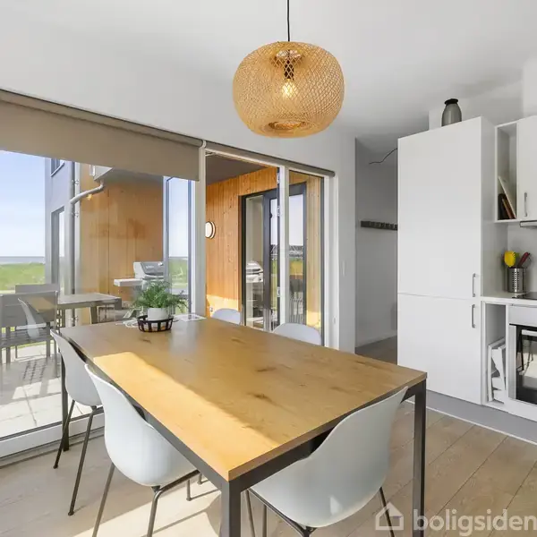 Spisebord med fire stole står i et lyst køkken med store glasdøre, der fører ud til en terrasse med udsigt over grønne marker. Lampe hænger over bordet.